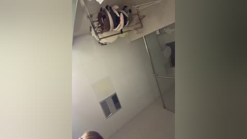 A video of a small, dimly lit room with a white ceiling, a beige wall, and a partially visible person in a striped shirt, standing near a shelf. The room appears sparse and utilitarian.