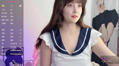 A video of a young East Asian woman with long brown hair, wearing a white sailor-style school uniform with a deep neckline, captured in a dimly lit room with a computer screen displaying a live stream.