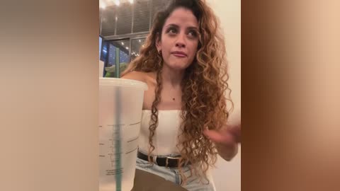 Video of a woman with curly, long brown hair, wearing a white off-shoulder top and high-waisted jeans, holding a plastic cup, standing in a dimly lit room with a visible ceiling fan.