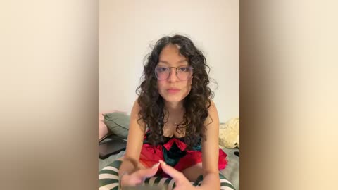 A video of a young woman with curly black hair, wearing glasses and a red and black dress, lying on a striped bed, smiling.