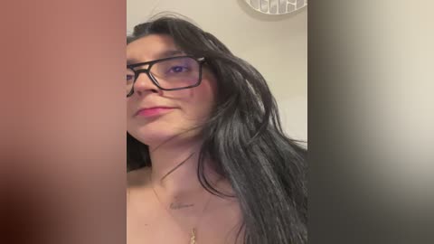 Media: A video of a woman with long black hair, glasses, and light skin, wearing a gold necklace. She has a tattoo on her chest. The background is blurred with a hint of beige and white.