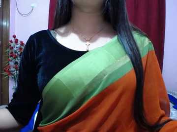 Media: Video of a woman in a black and orange sari with green and yellow accents, wearing a gold necklace. Background includes red curtains and a floral arrangement.