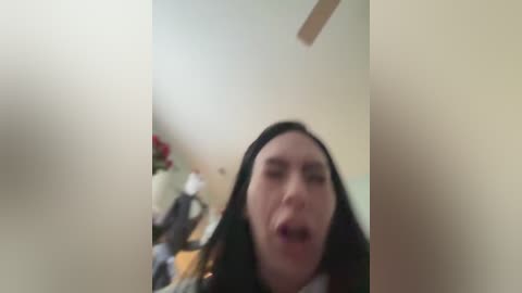 Media: Video of a young woman with long black hair, blurred face, in a room with beige walls, cluttered with items and a ceiling fan.