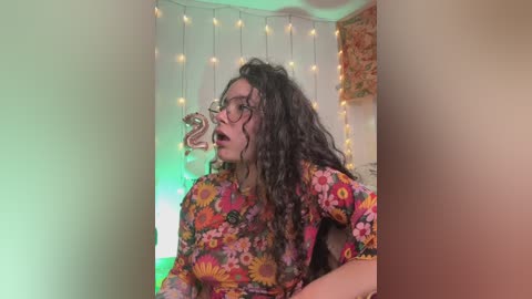 Video of a woman with curly dark hair, wearing glasses, a colorful floral shirt, and a large pink balloon with \"2\" in a cozy room with fairy lights.