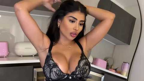 Video of a curvaceous, light-skinned woman with long black hair, wearing a black lace bra, posing in a modern kitchen with white cabinets and pink appliances.