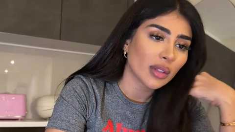 Video of a young woman with long black hair, wearing a grey shirt, speaking with a neutral expression, in a modern kitchen with beige tiles and pink items in the background.