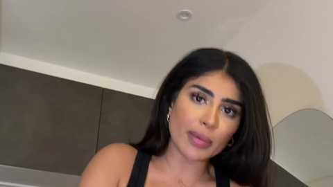 Video of a Latina woman with long black hair, wearing black clothing, and light makeup, smiling slightly, in a modern room with gray walls and a beige lamp.