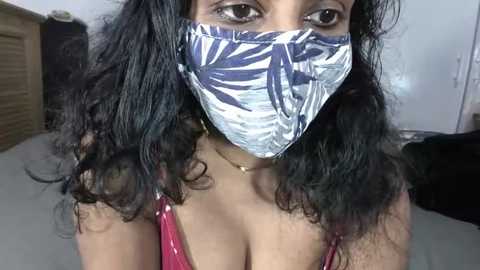 Media: Video of a young woman with medium brown skin, wearing a blue and white leaf-patterned face mask, revealing her dark brown eyes and curly black hair. She has a gold necklace and wears a pink spaghetti-strap top, indoors with a wooden door and white wall background.