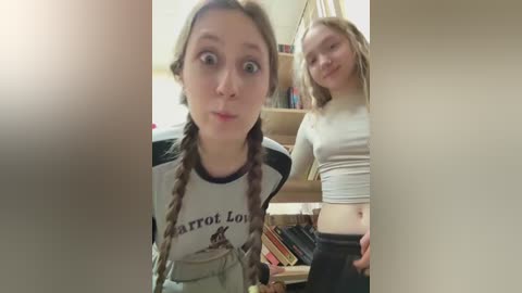 Video of two teenage girls in casual outfits, one with braided hair, the other with long hair, standing in a cluttered room with books and a mirror.