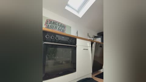A video of a minimalist, modern kitchen featuring a built-in black oven and microwave, a wooden countertop, and a small skylight window, with a \"Kangar\" sign on the wall.