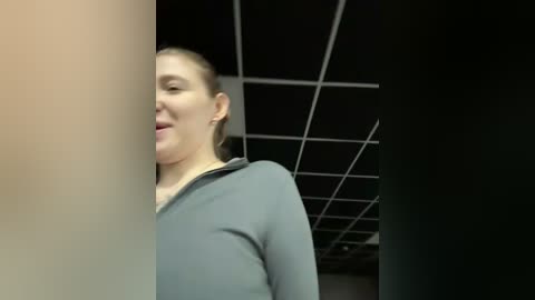 Video of a woman with light skin, brown hair tied back, wearing a gray shirt, standing in a dimly lit office with black ceiling tiles and fluorescent lights.