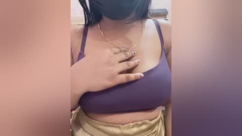 Video of a woman with medium-dark skin, wearing a purple sports bra, beige shorts, and a black face mask, covering her mouth. Her left hand rests on her chest, and she wears a gold necklace.