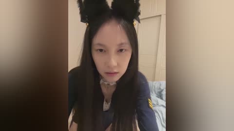 Video of an Asian woman with long black hair, wearing a navy-blue police uniform with a badge, cat ears, and a white collar. She sits on a bed in a dimly lit bedroom with a white wardrobe and blue sheets.