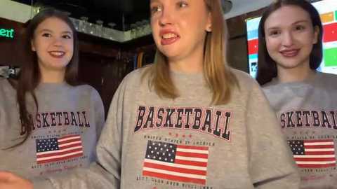 Video of three young women with long hair in basketball-themed gray t-shirts, smiling in a dimly-lit bar.
