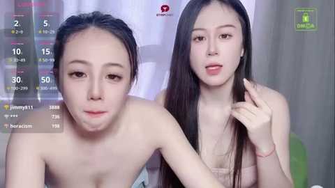 Video of two young Asian women with light skin, long dark hair, and slender figures. Both are topless, one with a white top, the other in a pink top. Background shows a white curtain.