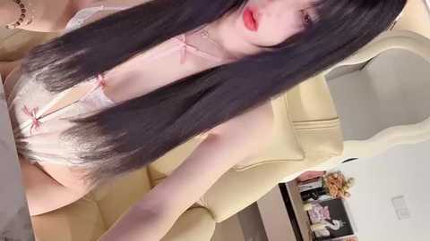 Video of an Asian woman with long, straight black hair, wearing a white lace lingerie bodysuit with a pink bow, seated on a beige leather chair in a modern, minimalistic room.