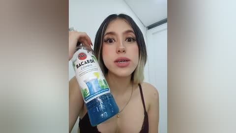 Video of a young, light-skinned Asian woman with long, dark hair and a slender build, wearing a black spaghetti strap top, holding a Bacardi bottle. She has a neutral expression.
