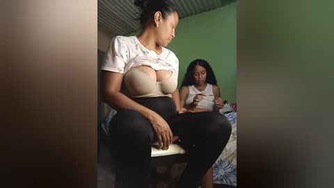 Video of a pregnant woman in a white shirt, exposing her large breasts, sitting beside a young girl with long hair in a green room.