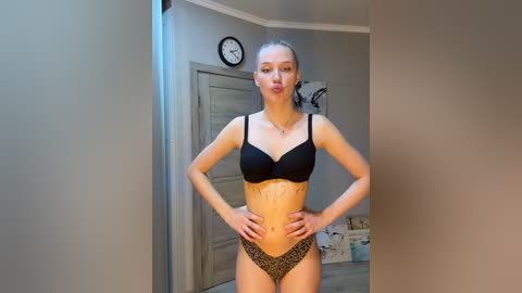 Media: Video of a slim, fair-skinned woman with long blonde hair in a ponytail, wearing black bra and leopard-print panties, standing confidently with hands on hips in a modern, neutral-toned bedroom.