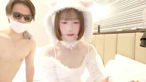 A video of an Asian man wearing sunglasses and a mask, standing next to a young Asian woman in a fluffy white bear costume, in a hotel room with blinds.