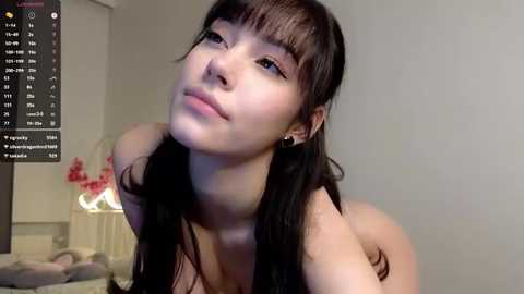 Video of an East Asian woman with straight black hair and light skin, wearing a black top, looking upward with a neutral expression, in a softly lit bedroom with pink flowers and a TV screen displaying an interface.