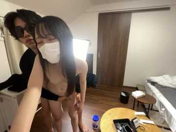 Video of two Asian women in a hotel room, one with long black hair, wearing a black bra and white mask, the other with short hair, also masked. Room is cluttered with luggage, clothes, and a bed.