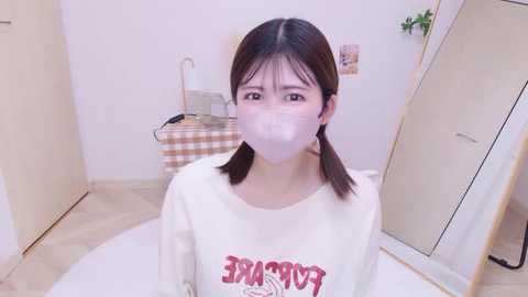 Video of an East Asian woman with straight black hair in pigtails, wearing a white long-sleeve shirt with red text, a white face mask, and standing in a clean, beige-tiled bathroom with a checkered laundry basket.
