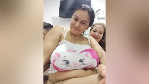 Video of a smiling woman with medium skin tone and dark hair, wearing a white tank top with a pink cat design, in a room with a TV and another woman in the background.