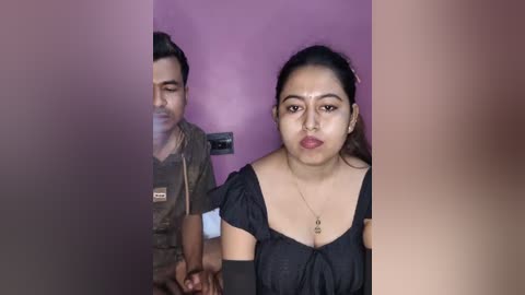 Video of a young South Asian woman with fair skin, dark hair, wearing a black top, standing in front of a purple wall, next to a man with a beard, in a dimly lit room.