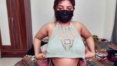 Video of a young woman with medium skin tone and dark hair, wearing a black face mask and a light green crochet top, standing in a cluttered bedroom with wooden furniture and a messy floor.