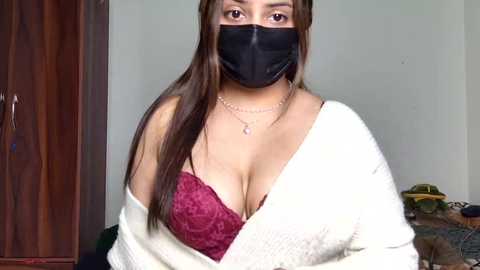 Video of a light-skinned woman with long brown hair, wearing a black face mask, maroon lace bra, and white cardigan, in a simple indoor setting with a wooden door and a cluttered background.