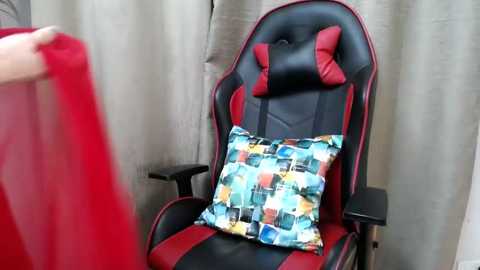Video of a person sitting in a red and black gaming chair with a colorful, patterned cushion, with a red cloth in the foreground.
