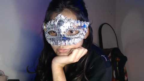 Video of a woman with long dark hair wearing a glittery masquerade mask and black dress, resting her chin on her hand, in a dimly lit room with a mirror and colorful clothing in the background.