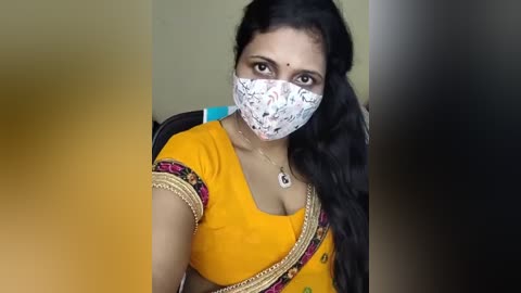 A video of a South Asian woman with medium skin tone and long, wavy black hair. She wears a yellow sari with intricate golden embroidery, a white floral mask, and a blue necklace.