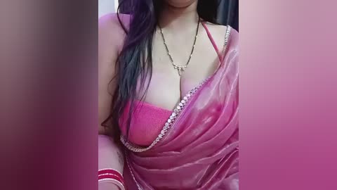 A close-up video of a woman with light skin, wearing a pink sari with a red lace bra visible, adorned with gold jewelry.