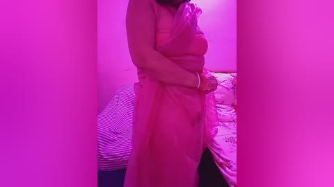 A video of a woman in a sheer, pink dress standing in a dimly lit room with pink lighting. She is partially hidden behind a curtain, and the background features a bed with a patterned quilt.
