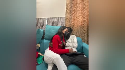 A video shows two Black women hugging on a teal couch; one in a red sweater, the other in a white sweater. The background features textured beige and grey walls, and a beige curtain.