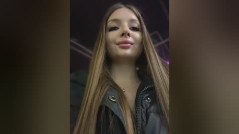 Video of a young woman with long, straight, light brown hair, wearing a black leather jacket, smiling slightly, with a blurred, dark background.