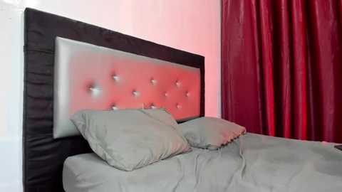 Video of a modern bedroom with a tufted, silver-gray upholstered headboard, two pillows, and gray bedding. The background features a maroon curtain and soft pink wall, creating a cozy atmosphere.