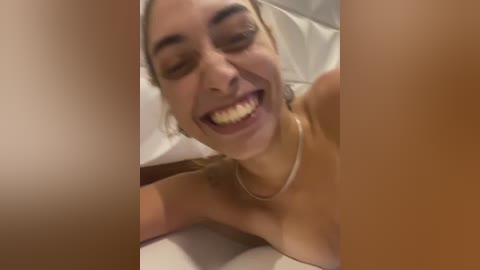 Video of a smiling, topless young woman with light skin, light brown hair, and a thin necklace, lying on a white bed. The image is framed by blurred, tan-toned areas on either side.
