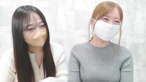 Video of two women with long black and blonde hair, wearing white surgical masks, white jackets, and gray sweaters, seated indoors with white walls and blurred furniture in the background.