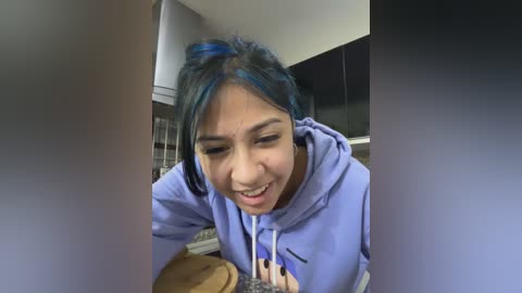 Media: A young woman with blue hair and a light purple hoodie leans over a table, smiling. She has medium skin tone and appears to be Asian. The background shows a kitchen with cabinets and a stove.