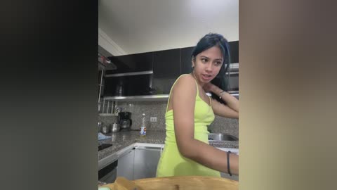 Media: Video of a young Asian woman with long black hair, wearing a neon yellow tank top, standing in a modern kitchen with stainless steel appliances and granite countertops.