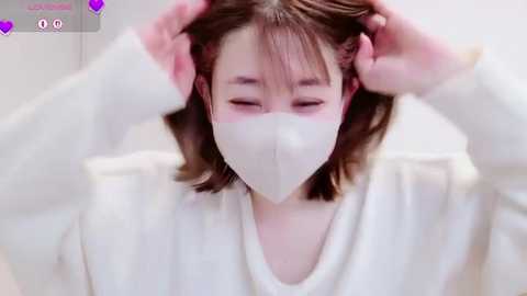 Media: Video of an Asian woman with light skin, wearing a white face mask, adjusting her shoulder-length brown hair with both hands. She has a calm expression.