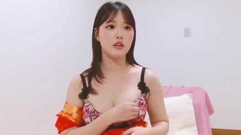 Video of an Asian woman with medium-length black hair, wearing a revealing red and black lace bra, standing in a minimalist bedroom with a pink bedspread.