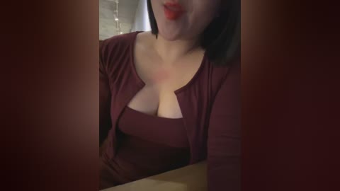 A video of a woman with fair skin and dark hair, wearing a deep burgundy top with a cutout neckline, licking her lips in a playful manner.