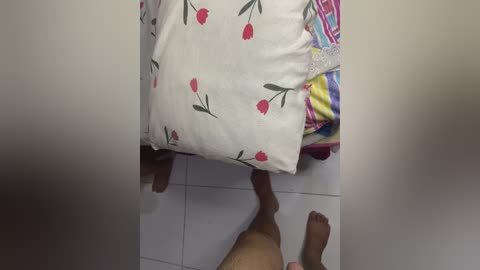 A video shows a person lying on a bed with white pillows and a colorful striped sheet, legs crossed, facing the camera. The room has white tiled flooring.