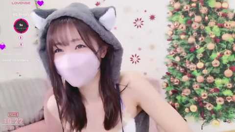 A young Asian woman with long black hair, wearing a grey cat hood and a pink mask, poses indoors with a decorated Christmas tree in the background.