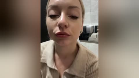 Video of a close-up, neutral expression Caucasian woman with fair skin, straight blonde hair, wearing a beige ribbed knit top. Background features a tiled wall with a paper towel dispenser and a trash bin.