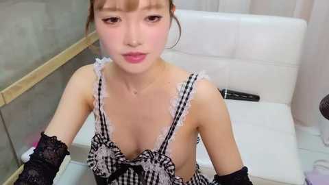 Video of an East Asian woman with light skin, brown hair in braids, wearing a black and white checkered maid outfit with lace details. She sits on a white chair in a modern bathroom, holding a black object.